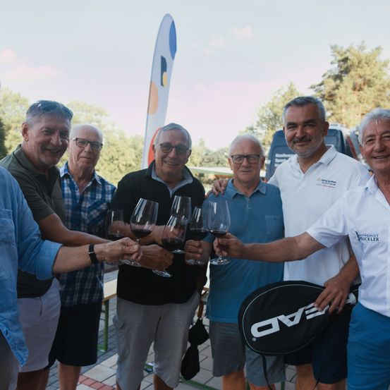 Eine Gruppe älterer Männer stoßen mit Weinläsern draußen an und lächeln. Ein Mann hält einen Tennisschläger. Sie sind in einem Park mit Bäumen und blauem Himmel.