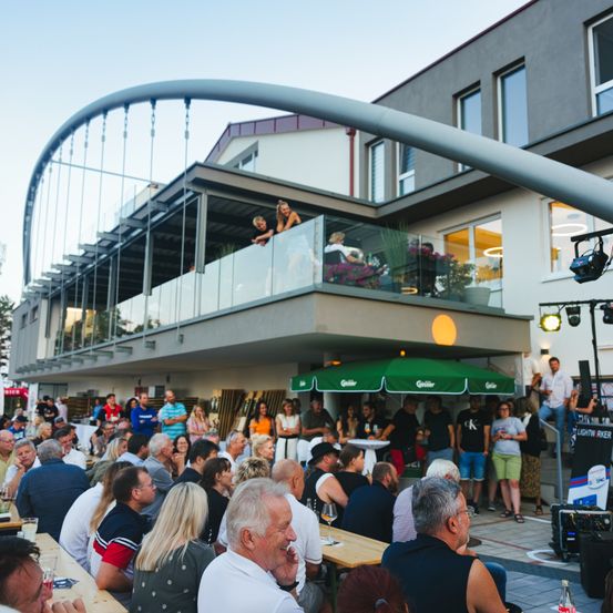 Eine Gruppe von Menschen versammelt sich im Freien für eine Veranstaltung. Sie sitzen um Tische herum unter einem Baldachin. Oben stehen zwei Personen auf einem Balkon, eine lehnt sich an das Geländer. Hinter ihnen steht ein Gebäude mit Glasfenstern und moderner Architektur.