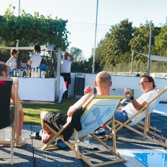 Eine Gruppe von Menschen entspannt sich auf Liegestühlen in der Nähe eines Pools. Ein Mann trägt ein Shirt mit der Nummer 43. Hinter ihnen steht ein Tisch mit Flaschen und Essen, und ein Mann steht daneben.