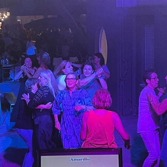 Eine Gruppe von Menschen tanzt in einem schwach beleuchteten Raum mit lila Lichtern. Ein Monitor mit der Aufschrift 'Amarillo' ist im Vordergrund zu sehen.
