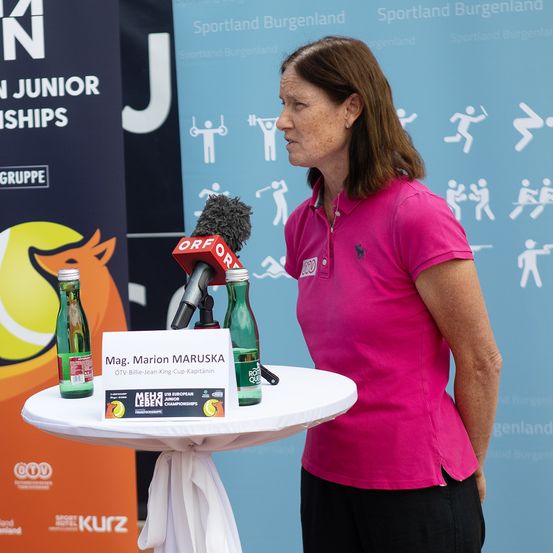 Ein Mann und eine Frau stehen vor einem Banner für die U18 European Junior Championships. Die Frau spricht in ein Mikrofon, und ein Tisch hält zwei Flaschen und ein Schild mit dem Namen Mag. Marion Maruschka.