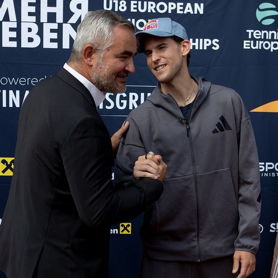 Drei Männer stehen gemeinsam auf einer Bühne mit einem blauen Hintergrund. Ein Mann in einem schwarzen Anzug schüttelt die Hand eines Mannes in einer grauen Kapuzenjacke. Ein weiterer Mann in einem grauen T-Shirt steht in der Nähe.