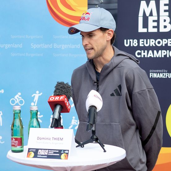 Dominic Thiem spricht an einem Podium mit Mikrofonen und zwei Flaschen, trägt eine Kappe und eine graue Adidas-Hoodie. Hinter ihm steht ein blauer Banner, auf dem 'Mehr Leben U18 European Junior Championships' steht. Ein anderer Mann in einem weißen T-Shirt steht in der Nähe.