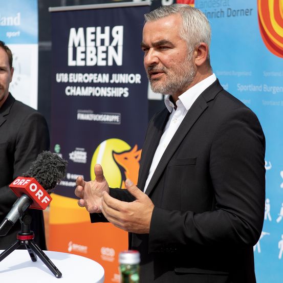 Ein Mann in einem schwarzen Anzug spricht in ein Mikrofon, möglicherweise bei einer Sportveranstaltung. Hinter ihm befindet sich ein blauer Banner mit weißem Text.