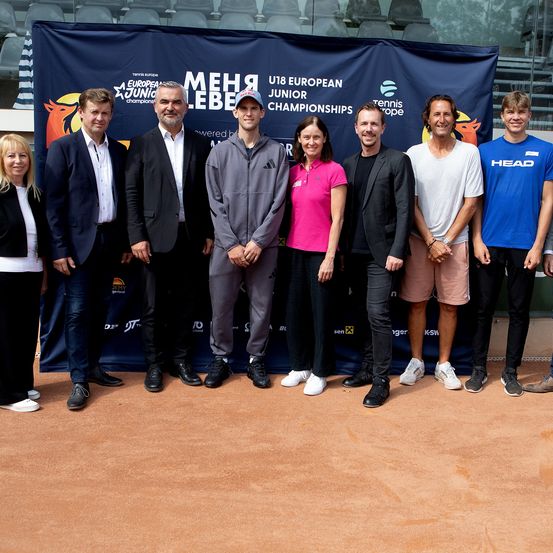 Eine Gruppe von Personen, einschließlich Männer und Frauen, steht auf einem Tennisplatz vor einem blauen Banner mit weißem Text. Sie scheinen sich bei einer Veranstaltung zu befinden.