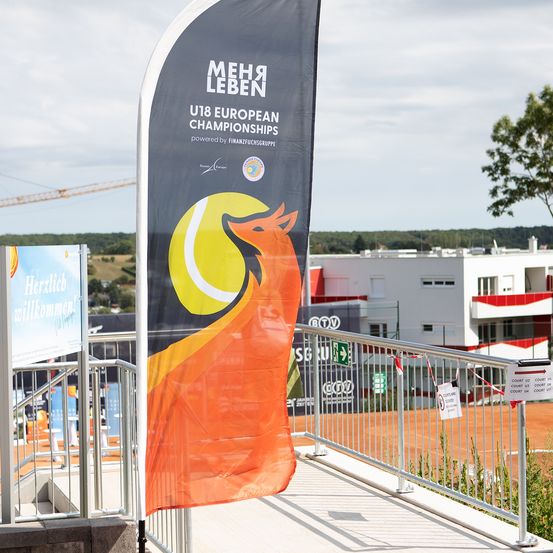 Ein Banner für die U18-Europameisterschaft mit einem Tennisball und einem Fuchs steht auf einer Metallschiene mit einem Tennisplatz im Hintergrund.