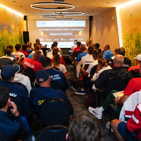 Eine Gruppe von Personen in Sporttrikots und Mützen sitzt in Stühlen in einem Raum. Eine Projektionsleinwand zeigt den Text 'Mehleben bei den Europäischen Juniorenmeisterschaften'. Lautsprecher stehen links und eine Person spricht. Die Wände sind mit Wandgemälden verziert.