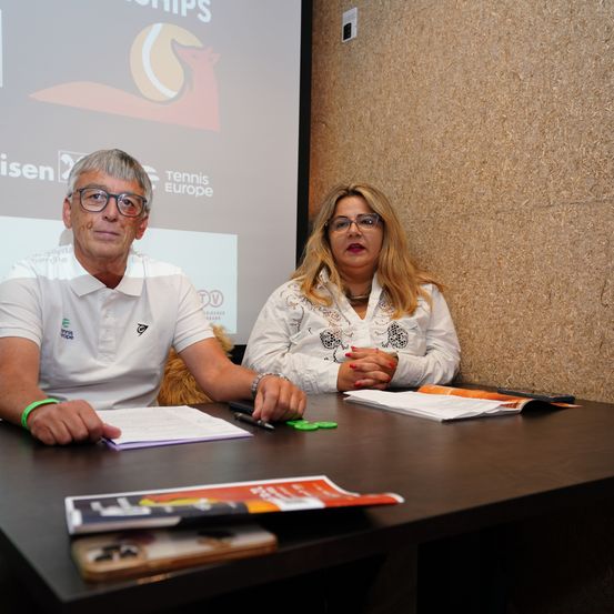 Zwei Personen sitzen an einem Tisch mit Papieren und Stiften vor einer Projektionsleinwand, die einen Tennisball und Text zeigt.