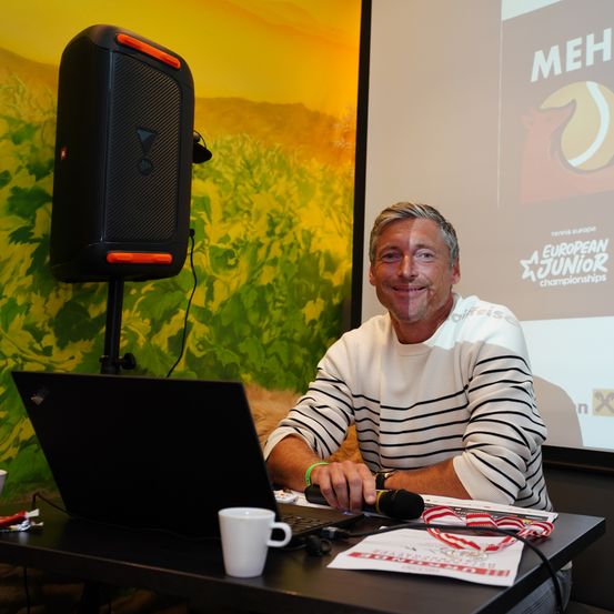 Ein Mann sitzt an einem Schreibtisch mit Laptop und Mikrofon, lächelt, mit einem Lautsprecher dahinter. Eine Projektionsleinwand zeigt ein Tennislogo.