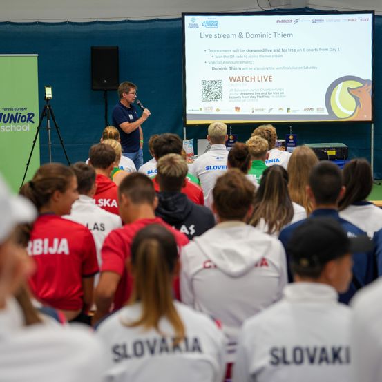 Ein Mann spricht in ein Mikrofon vor einer Projektionsleinwand. Das Publikum besteht aus jungen Menschen in roten und weißen Shirts. Die Leinwand zeigt einen Live-Stream und Dominic Thiem.