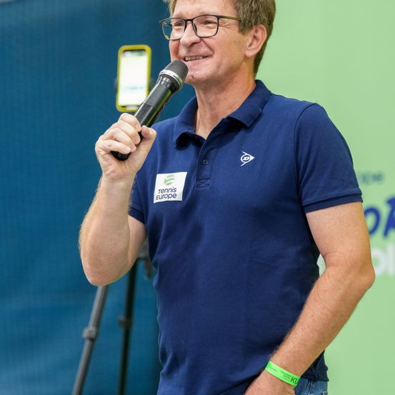 Ein Mann spricht in ein Mikrofon und trägt ein blaues Polohemd mit einem weißen Tennis Europe-Abzeichen und einem grünen Armband.