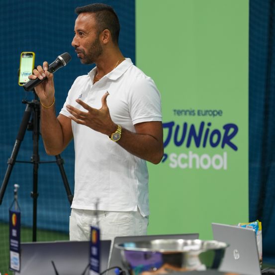 Ein Mann in einem weißen Poloshirt und einer Uhr spricht in ein Mikrofon vor einer grünen Wand mit den Worten Tennis Europe Junior School.