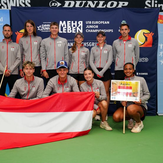 Eine Gruppe von Tennisspielern in grauen Jacken posiert für ein Foto mit einer österreichischen Flagge vor einem blauen Hintergrund mit dem Text 'U18 European Junior Championships.'
