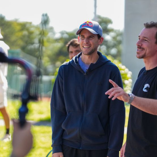 Zwei Männer in Freizeitkleidung stehen in einem Park, einer lächelt. Hinter ihnen steht eine Person in der Nähe eines Zauns, und ein verschwommenes Bild eines Gebäudes und Bäume ist sichtbar.