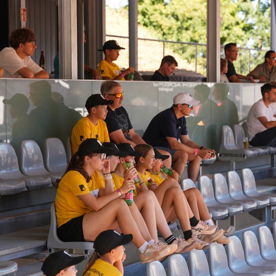 Eine Gruppe von Menschen in gelben T-Shirts sitzt auf Tribünen und trinkt aus grünen Flaschen. Hinter ihnen sitzen andere im Hintergrund, einige tragen Hüte. Die Umgebung ist im Freien, mit Bäumen in der Ferne.
