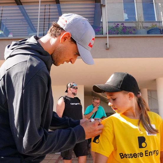 Ein Mann in einer Red Bull Kappe befestigt einen Abzeichen an einem Mädchen, das ein gelbes Shirt mit den Worten 'Bessere Enrg' trägt. Dahinter raucht ein anderer Mann.