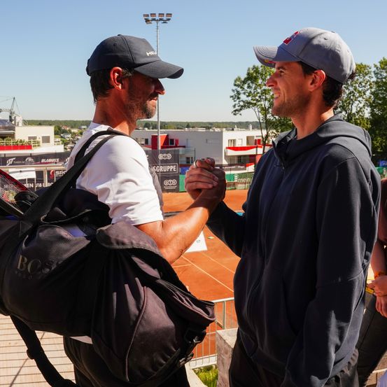 Zwei Männer schütteln sich die Hände auf einem Tennisplatz. Einer trägt einen schwarzen Hut, einen Rucksack und ein weißes T-Shirt. Der andere trägt einen grauen Hut und eine blaue Kapuzenjacke. Ein Tennisschläger und ein Tennisplatz sind im Hintergrund zu sehen.