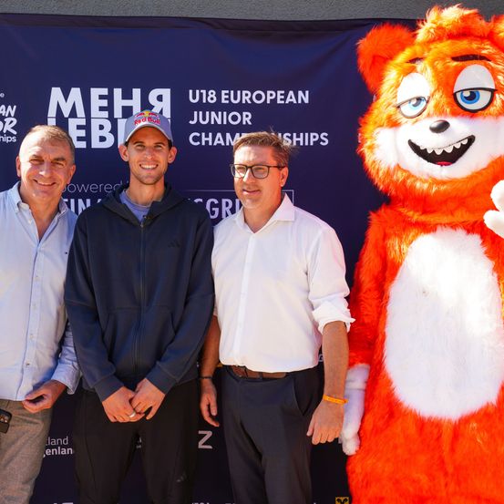 Drei Männer und eine Maskottchen stehen vor einem Banner, das die U18 European Junior Championships bewirbt. Die Maskottchen ist ein orange-weißer Charakter.