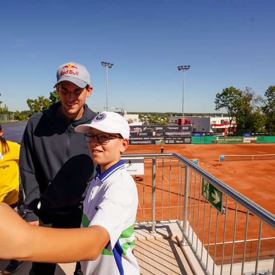 Ein Mann mit einer Red Bull Kappe und ein Junge in einem weißen Shirt posieren für ein Foto auf einem Tennisplatz mit einem Metallgeländer. Bäume und Gebäude sind im Hintergrund.
