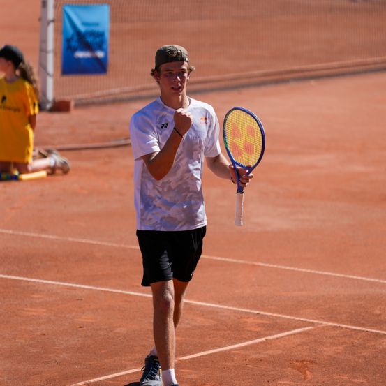 Ein junger Mann auf einem Tennisplatz, der ein weißes T-Shirt und schwarze Shorts trägt, hält einen Tennisschläger und scheint zu feiern.