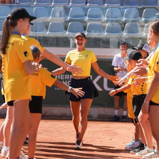 Eine Gruppe junger Mädchen in gelben Uniformen und schwarzen Shorts feuert eine Tennisspielerin auf einem Platz mit leeren blauen Stühlen im Hintergrund an.