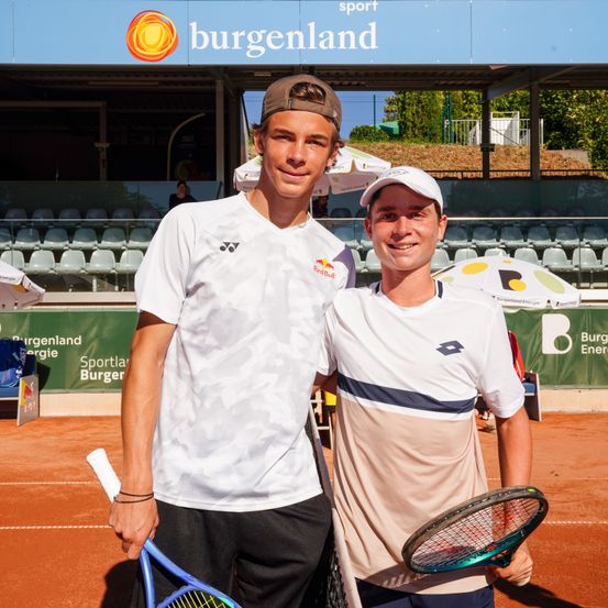 Zwei Männer stehen auf einem Tennisplatz mit Schlägern, in weißen Shirts. Hinter ihnen befindet sich ein leeres Stadion mit einem blauen Banner, das 'Burgenland Sport' liest.