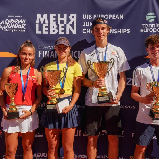 Vier Tennisspieler, zwei Frauen und zwei Männer, stehen zusammen und halten Pokale vor einem Banner, auf dem 'U18 European Junior Championships' steht. Sie tragen alle Medaillen um den Hals.