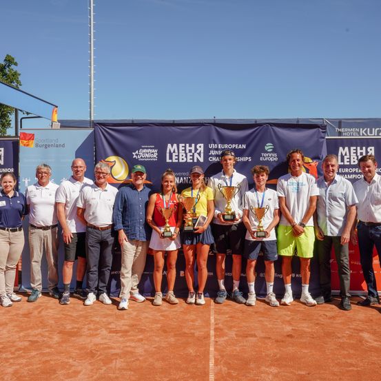 Eine Gruppe von Personen steht auf einem Tennisplatz und hält Trophäen. Sie sind von Bannern und einer Wand mit Logos umgeben. Die Gruppe besteht aus mehreren Männern und Frauen, einige tragen Hüte und Turnschuhe. Der Hintergrund zeigt Bäume und einen klaren Himmel.