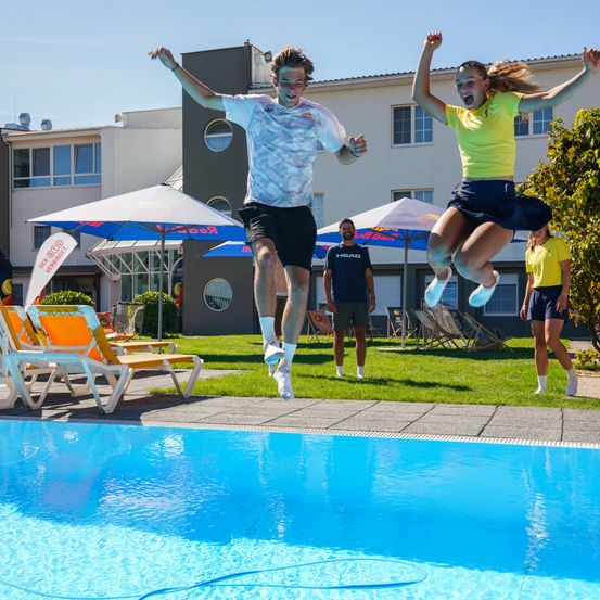 Zwei Personen springen in einen Pool, tragen Turnschuhe und Shorts. Hinter ihnen steht ein Gebäude mit Fenstern und Sonnenschirmen. Ein Mann steht in der Nähe und schaut sie an.