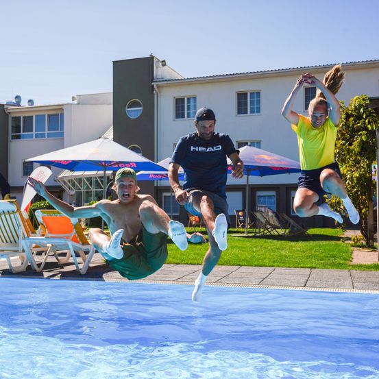 Drei Menschen springen fröhlich in einen Pool. Der Mann in der Mitte trägt ein blaues Shirt mit der Marke 'HEAD'. Zwei Personen tragen Turnschuhe. Ein Gebäude mit Fenstern steht hinter ihnen.