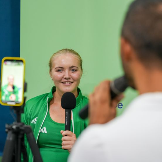 Eine Frau in einem grünen Jackett wird von einem Mann mit Mikrofon interviewt. Ein Handy auf einem Stativ hinter ihr nimmt das Interview auf.