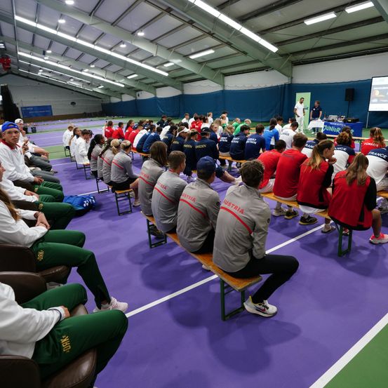 Eine Gruppe von Athleten aus verschiedenen Ländern in Sportkleidung sitzt auf Bänken in einer Sporthalle und hört möglicherweise eine Ankündigung.