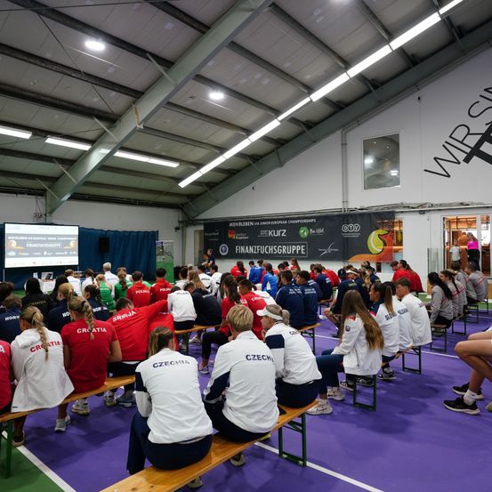 In einer überdachten Sporthalle sitzen Athleten aus verschiedenen Ländern auf Bänken. Sie tragen Uniformen mit unterschiedlichen Ländernamen. Eine Leinwand auf der linken Seite zeigt Informationen an. Banner und ein Logo sind an der Wand angebracht.