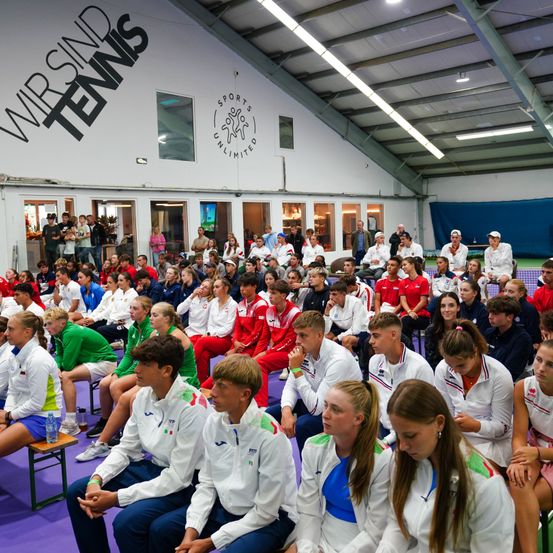 Eine große Gruppe von Athleten in Sportuniformen sitzt in einer Reihe, wahrscheinlich in einer Sporteinrichtung. Sie blicken nach vorne und warten möglicherweise auf den Beginn eines Spiels. Die Einrichtung hat eine weiße Wand mit den Worten 'Wir sind Tennis' und einem Logo. Im Hintergrund stehen Personen, vermutlich Zuschauer.