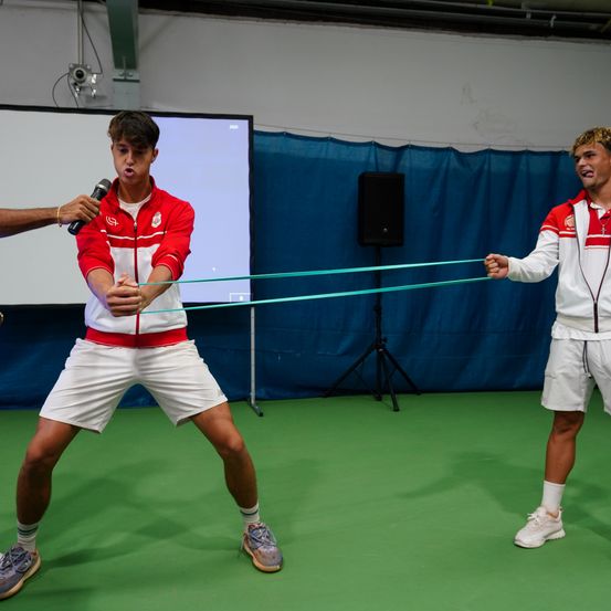 Zwei Athleten ziehen an einem grünen Band, während einer ein Mikrofon, einen Lautsprecher und eine Leinwand im Hintergrund hält.