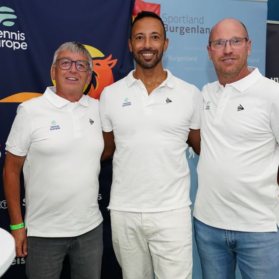 Drei Männer stehen eng beieinander vor einem Banner für Sportland Burgenland. Sie tragen alle weiße Polo-Shirts mit Logos. Der Mann auf der linken Seite hat ein grünes Armband.