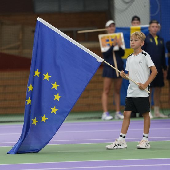 Ein junger Junge trägt eine europäische Flagge und geht auf einem Tennisplatz, mit anderen im Hintergrund.