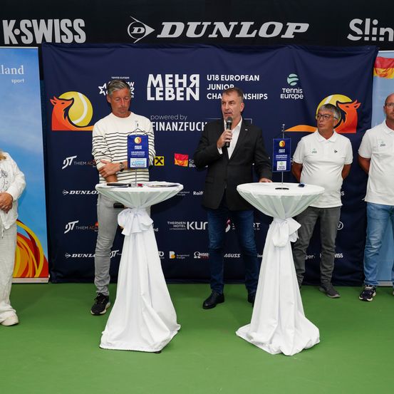 Fünf Männer stehen auf einem grünen Feld, einer mit einem Mikrofon, die ein Publikum ansprechen. Sie sind von weißen Tischen umgeben, die mit Schleifen geschmückt sind. Hinter ihnen befindet sich ein blauer Hintergrund mit dem Logo und Text einer Tennismeisterschaft.