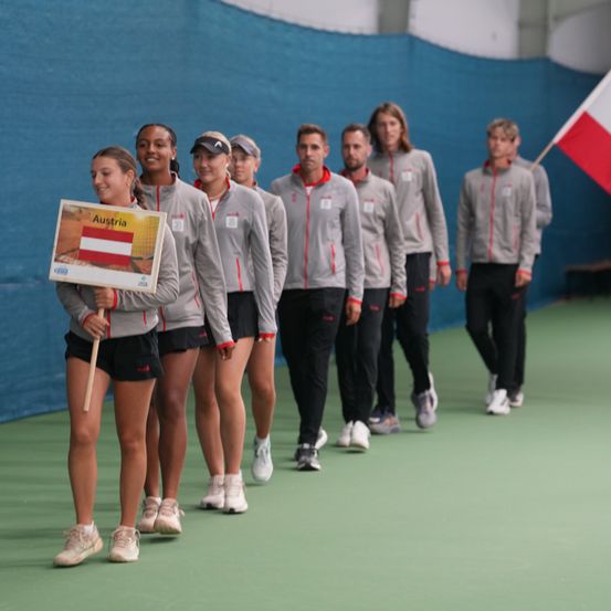 Eine Gruppe von Tennisspielern aus Österreich läuft auf einem Platz, mit einer Flagge im Hintergrund.