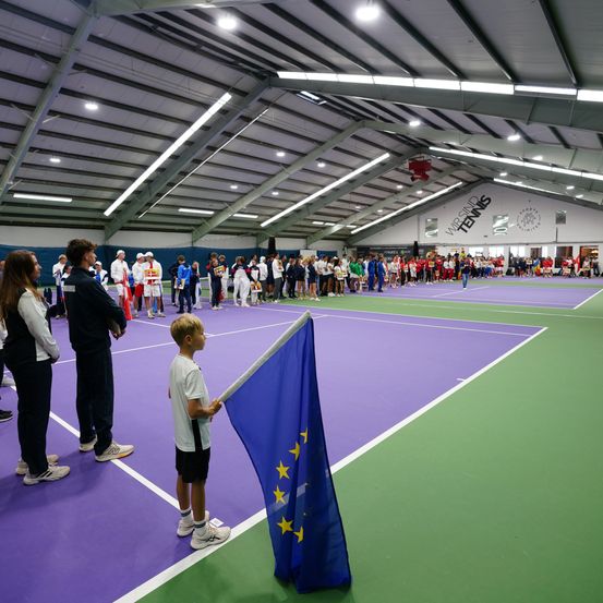 Überdachter Tennisplatz mit einem Jungen, der eine EU-Flagge hält. Zuschauer in Sportkleidung schauen zu, einige halten Flaggen. Der Platz hat eine lila-grüne Belagsfläche.