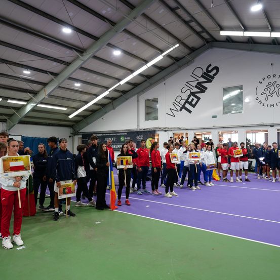 Eine Gruppe von Athleten steht auf einem violetten und grünen Tennisplatz und hält Schilder mit den Flaggen ihrer Länder. Sie sind in Sportuniformen gekleidet. Die Wand trägt den Text Wirsind Tennis und ein Logo.
