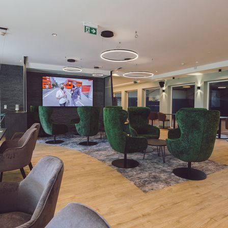 Ein Innenbereich mit Holzböden und mehreren grünen Drehsesseln. Ein großer Bildschirm zeigt ein Video, und es gibt runde Deckenleuchten.