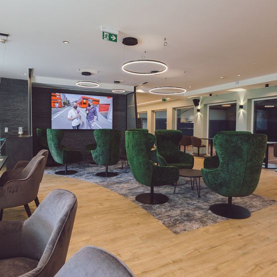 Ein Innenbereich mit Holzböden und mehreren grünen Drehsesseln. Ein großer Bildschirm zeigt ein Video, und es gibt runde Deckenleuchten.