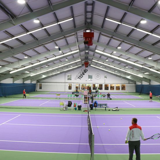 Ein Hallentennisplatz mit Spielern auf dem Platz. Der Platz hat eine lila Oberfläche. Die Decke wird von Metallträgern gestützt. Tennisbälle sind auf dem Platz verstreut.