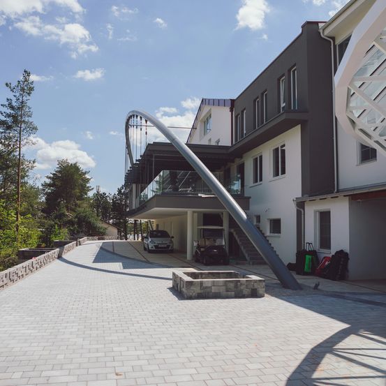 Ein modernes architektonisches Design zeigt einen großen gewölbten Eingang mit einem geparkten Auto. Der Einfahrtsweg führt zu einem Carport, wo ein Golfwagen geparkt ist. Die Szene ist vor dem Hintergrund von Bäumen und einem blauen Himmel angesiedelt.