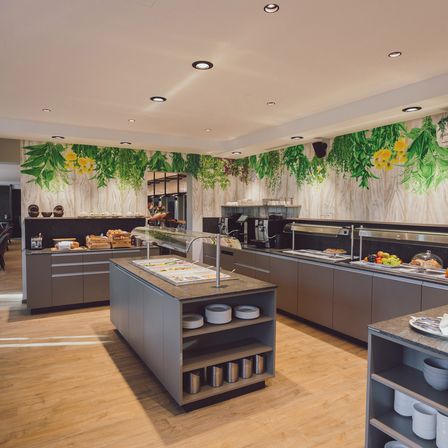 Ein moderner, gut beleuchteter Küchenbereich mit Holzböden, grauen Schränken und einem Buffettisch mit einer Vielzahl von Speisen. Die Wände sind mit grünen Blättern und gelben Blumen dekoriert.