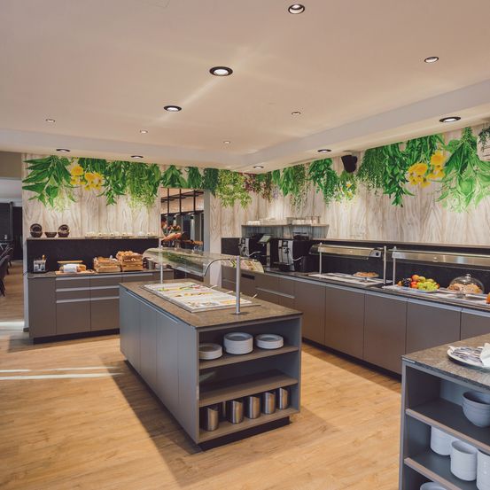 Ein moderner, gut beleuchteter Küchenbereich mit Holzböden, grauen Schränken und einem Buffettisch mit einer Vielzahl von Speisen. Die Wände sind mit grünen Blättern und gelben Blumen dekoriert.