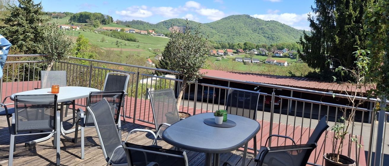 Ein Balkon mit Tischen und Stühlen blickt auf ein Tal mit einem Berg in der Ferne unter einem blauen Himmel mit einigen Wolken.