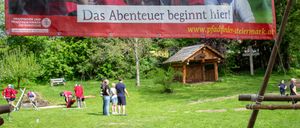 Eine Gruppe von Menschen befindet sich in einem Park mit einem großen Banner, auf dem steht: 'Das Abenteuer beginnt hier!' Einige Leute stehen, während andere sich bücken und etwas tun.