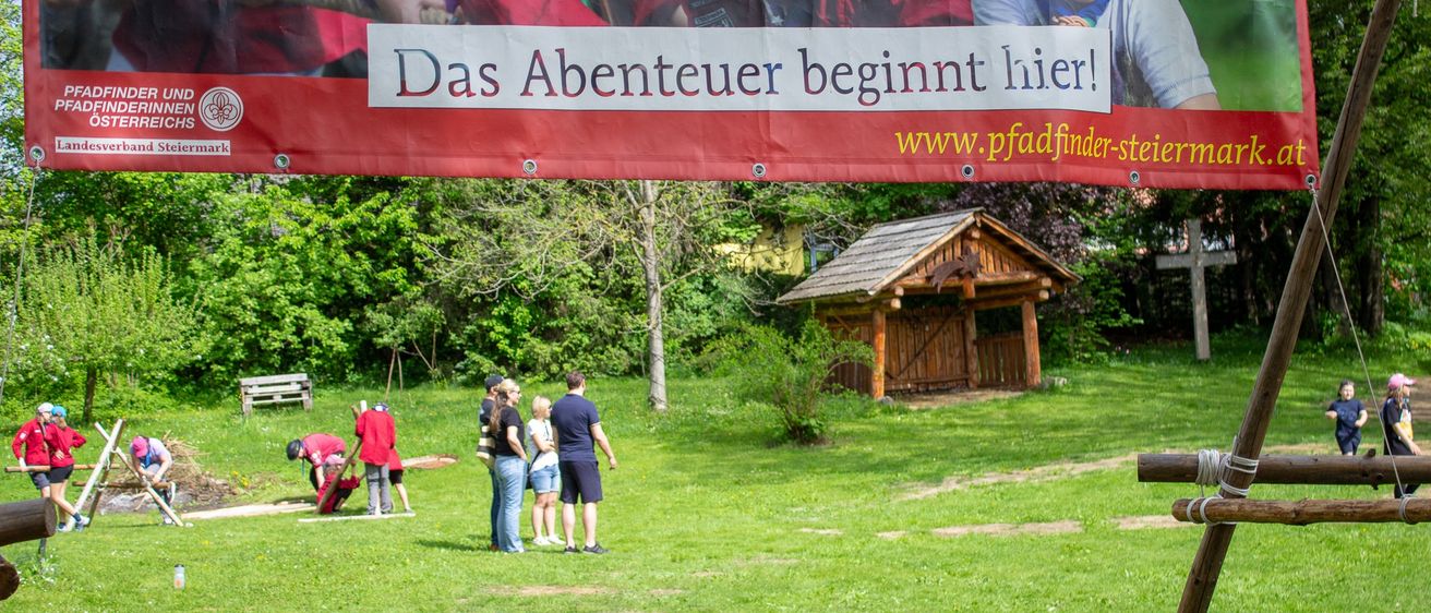 Eine Gruppe von Menschen befindet sich in einem Park mit einem großen Banner, auf dem steht: 'Das Abenteuer beginnt hier!' Einige Leute stehen, während andere sich bücken und etwas tun.